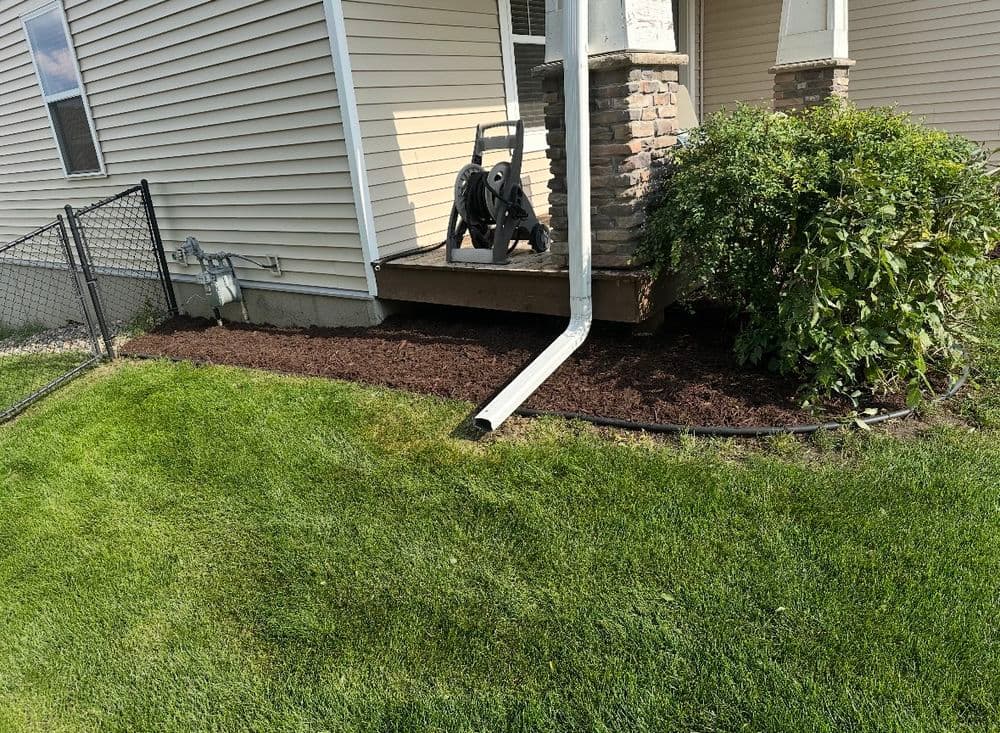 Mulched garden area beside a house with a downspout and green lawn in sunlight.