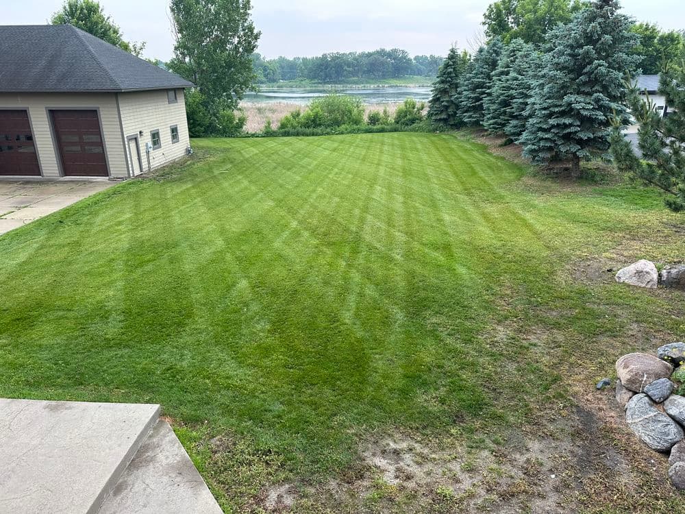 Lush, manicured lawn with striped patterns, surrounded by trees and a garage, near a tranquil lake.