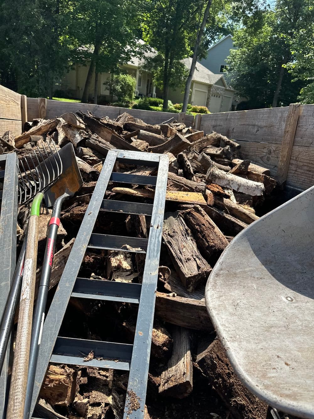 Lumber stacked in a trailer with gardening tools and a wheelbarrow in a lush backyard.