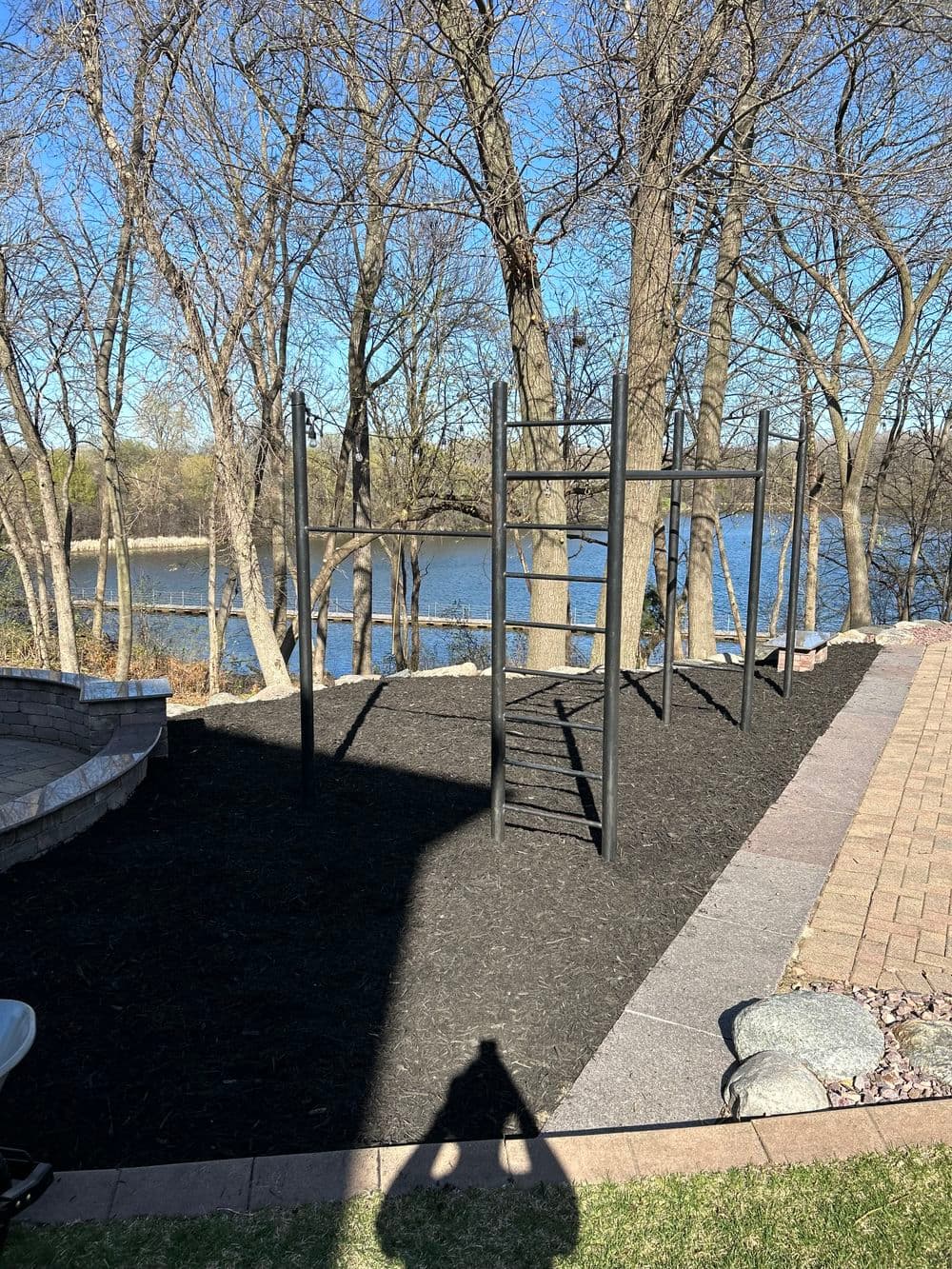 Outdoor playground with climbing bars beside a river, surrounded by bare trees and mulch.