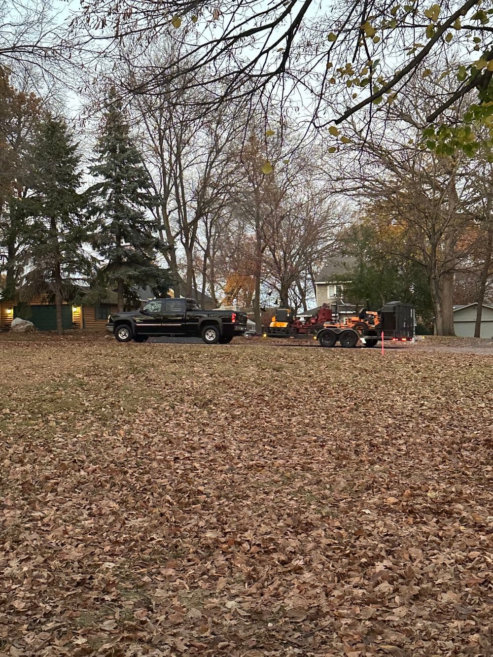 Pickup truck and trailer with equipment parked in a yard covered in autumn leaves.