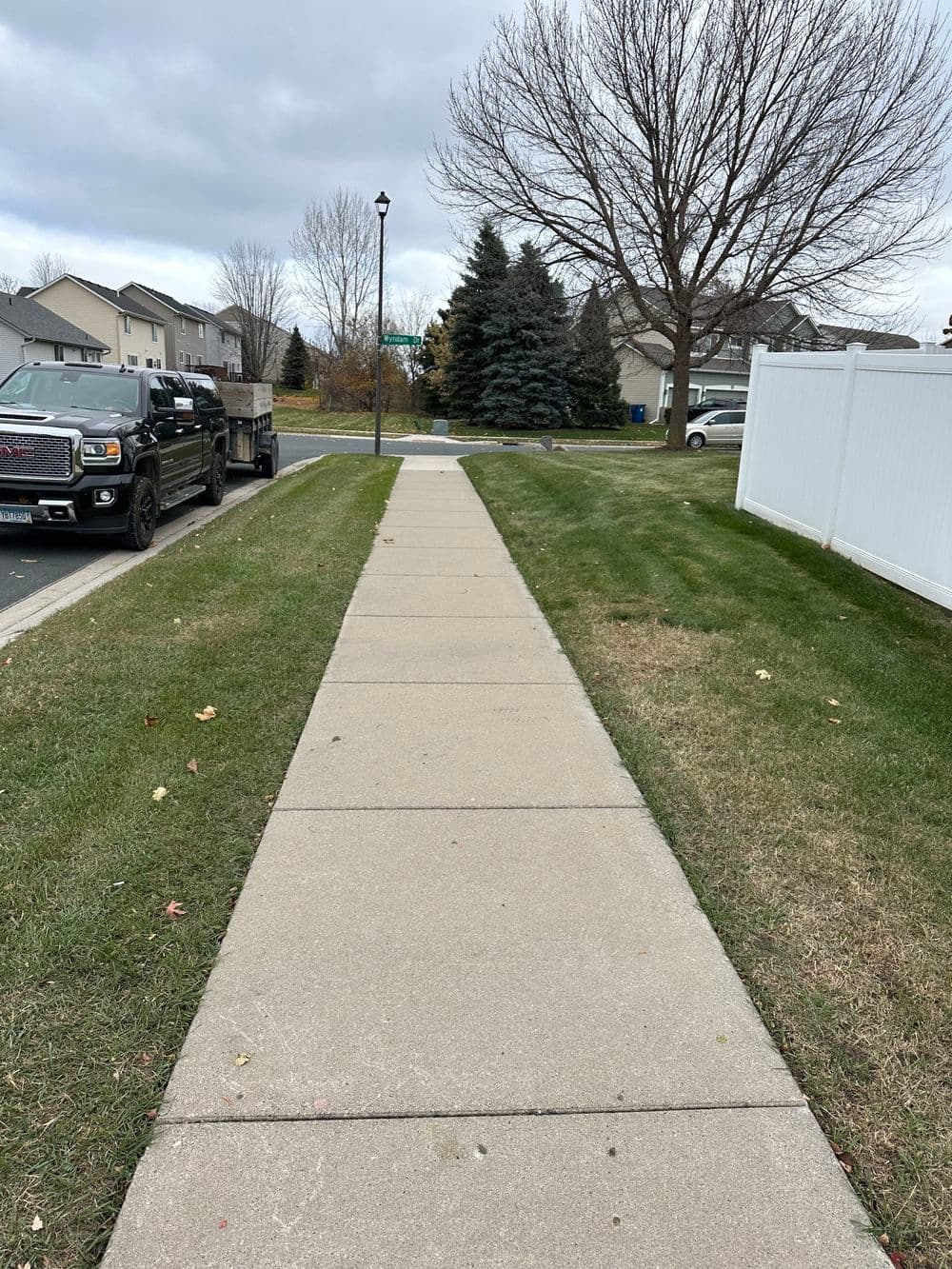 Concrete sidewalk pathway with grass and trees lined by houses and parked trucks.
