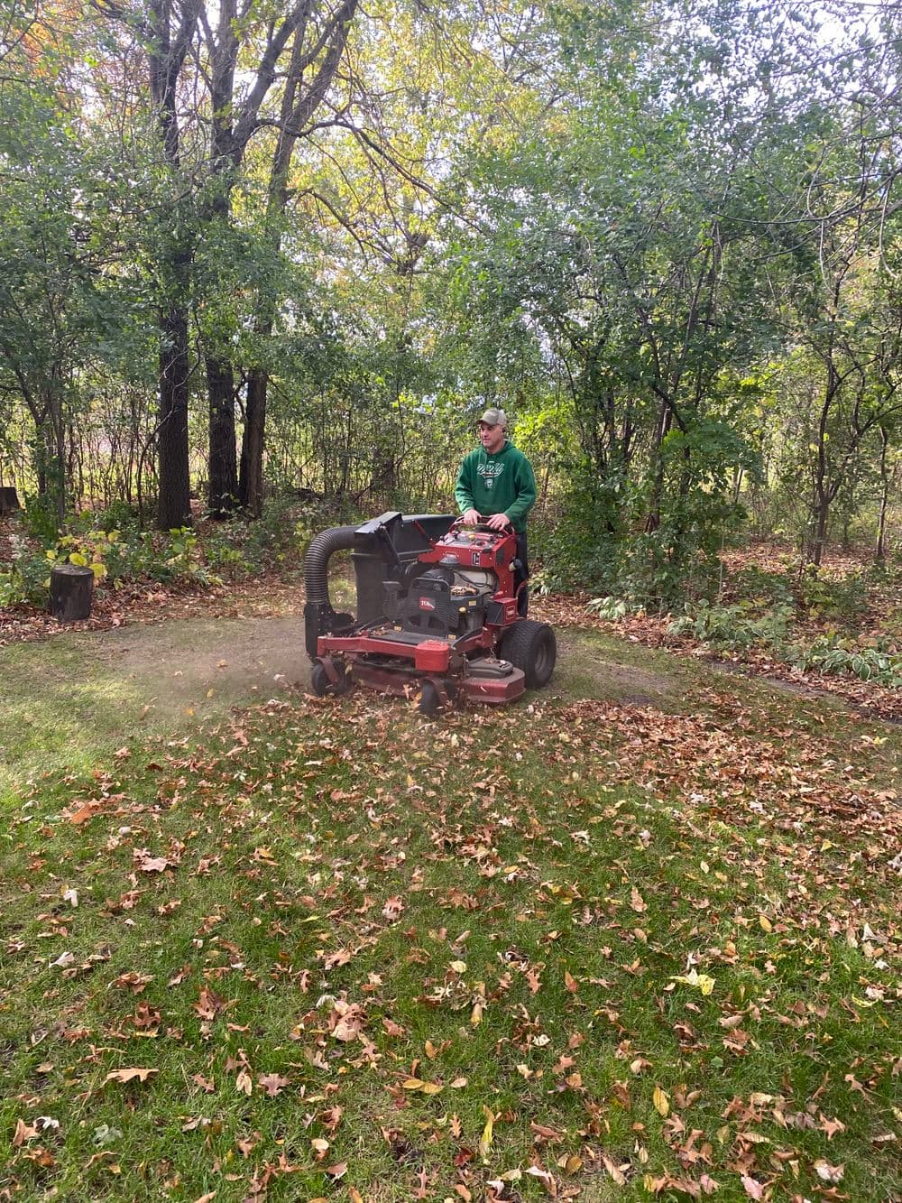Person using a lawn mower to clear fallen leaves in a wooded area during autumn.