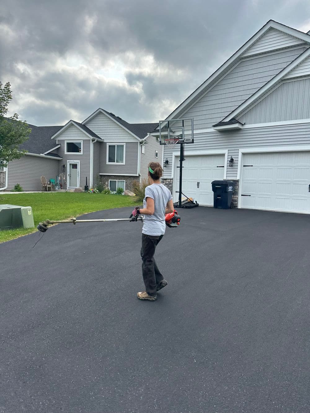 Person using a string trimmer on a freshly paved driveway near residential homes.