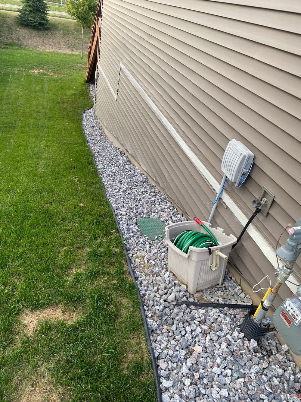 Gravel landscaping along a house with a water hose and utility access point.