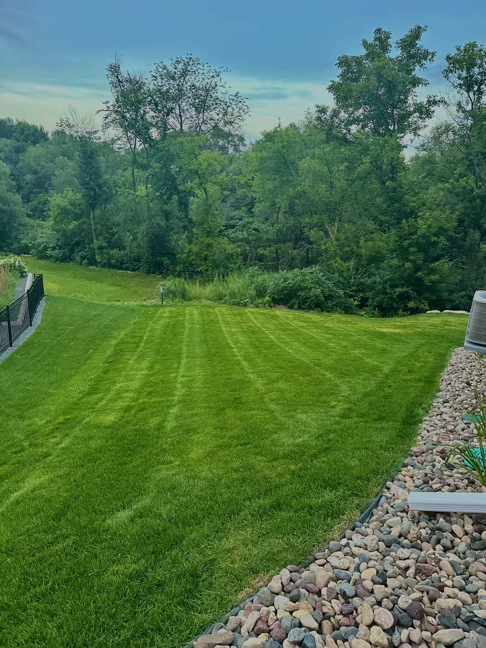 Lush green lawn with manicured stripes and wooded area in the background, featuring landscape stones.