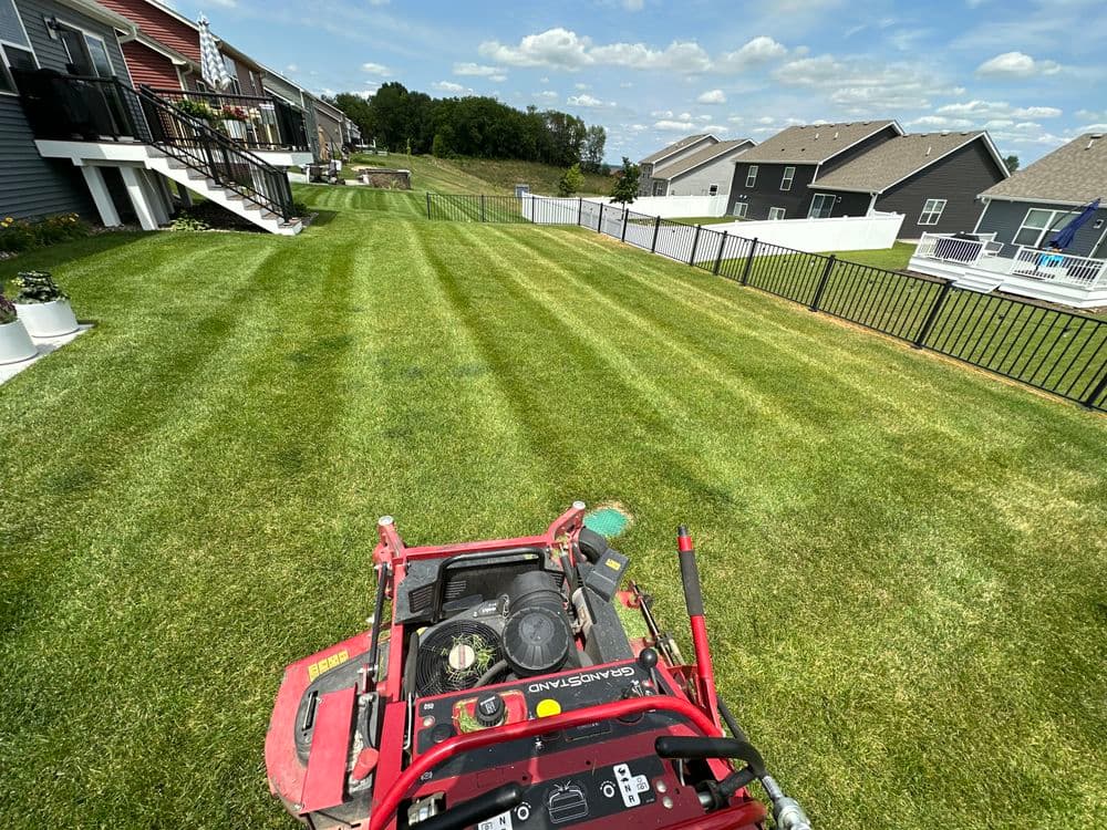 Lawn care scene showing a mower on freshly cut grass with neighboring houses in the background.