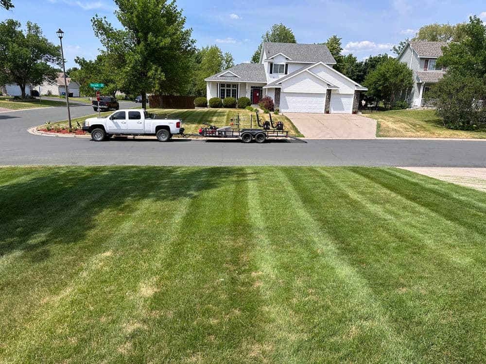White pickup truck towing a trailer parked on a suburban street with neatly striped green lawn.