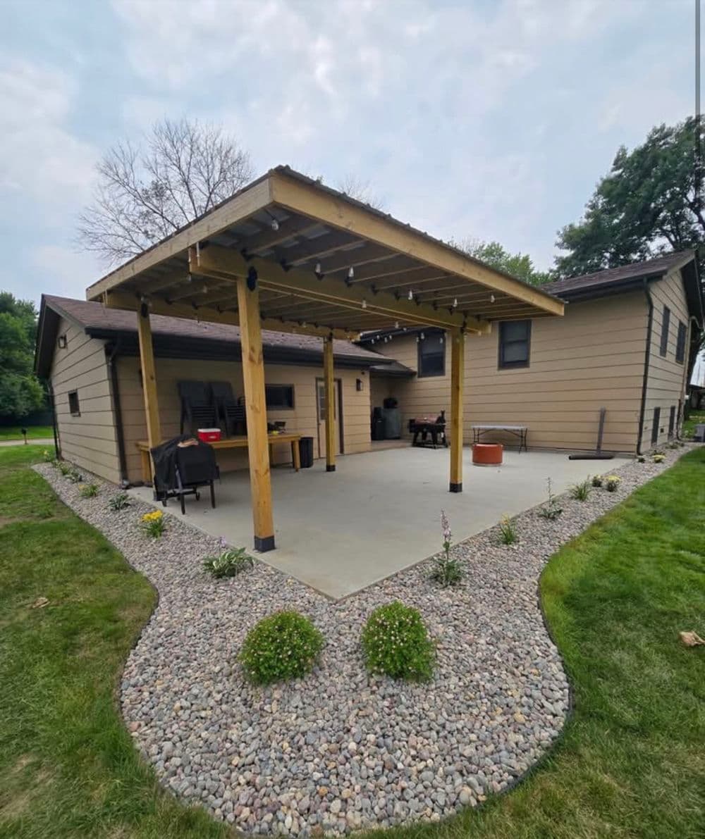A cedar pergola over a concrete patio with a curved river-rock border and foundation plantings