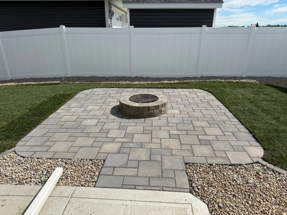 A rectangular paver patio with a round stacked-stone fire pit at its center, laid into fresh sod, with a gravel band along a white vinyl privacy fence and a dark-sided house behind
