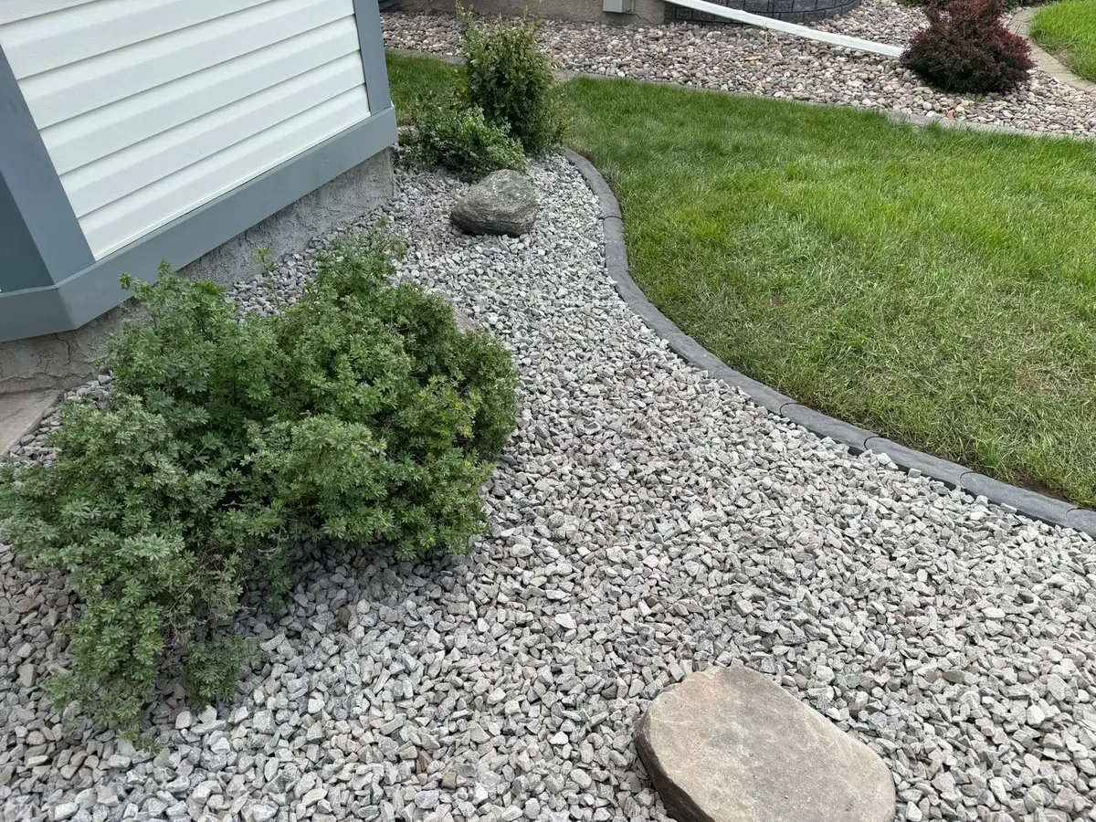 A bed of light grey river rock running along a pale blue-sided house, planted with low green shrubs and set with a flat flagstone step, held off the mown lawn by a curved dark grey edge