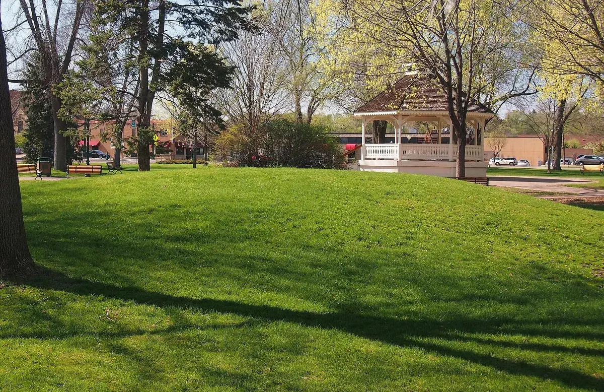 A white gazebo at the top of a broad mown lawn under mature shade trees at Chaska Public Square