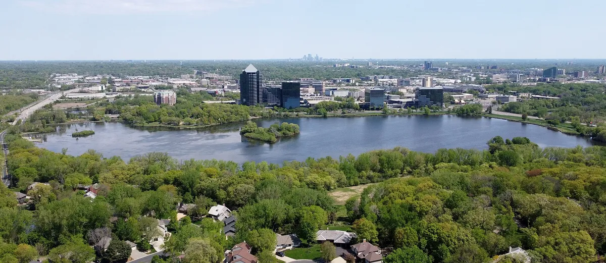 Normandale Lake seen from above, ringed by dense tree canopy with the Minneapolis skyline on the horizon