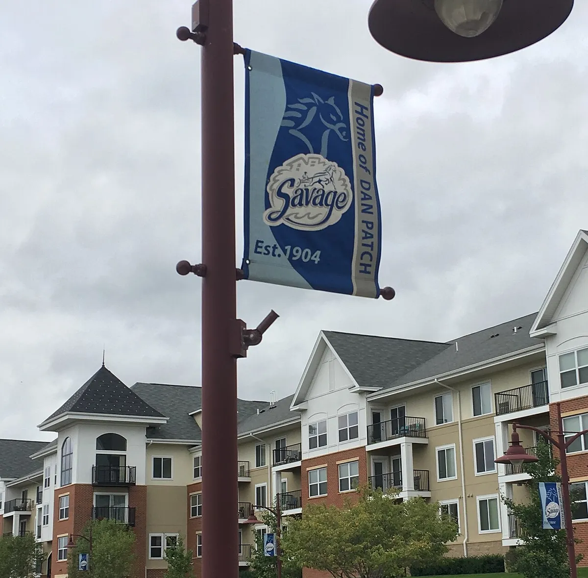 Street banners on a lamppost reading Savage, Est. 1904, Home of Dan Patch, with apartment buildings behind